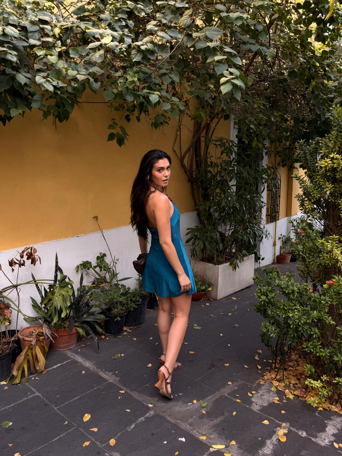 A woman wearing a teal blue sleeveless mini dress and tan high-heeled sandals stands on a stone walkway in a garden courtyard with a yellow wall, potted plants, and leafy vines overhead.